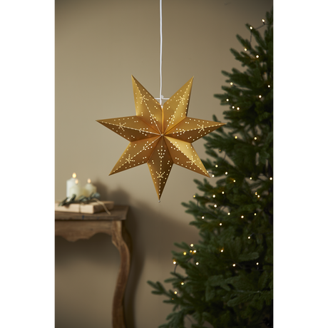 The Star Trading Classic Hanging Star Gold (16.5") glows near a Christmas tree with white lights. Candles and gifts on a wooden table add to the cozy holiday decor.
