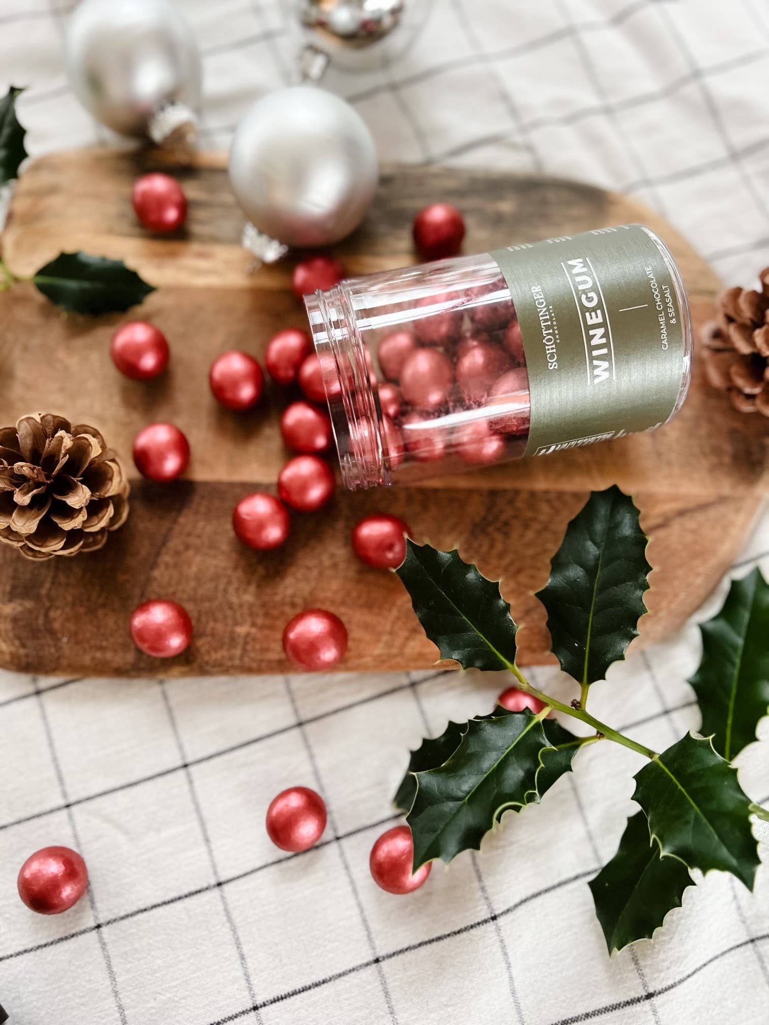 A jar opened to reveal the Classic Winegum Seasalt & Caramel Chocolate candies, rich in color like red amber, sits on a wooden board surrounded by silver ornaments, pinecones, and holly leaves.