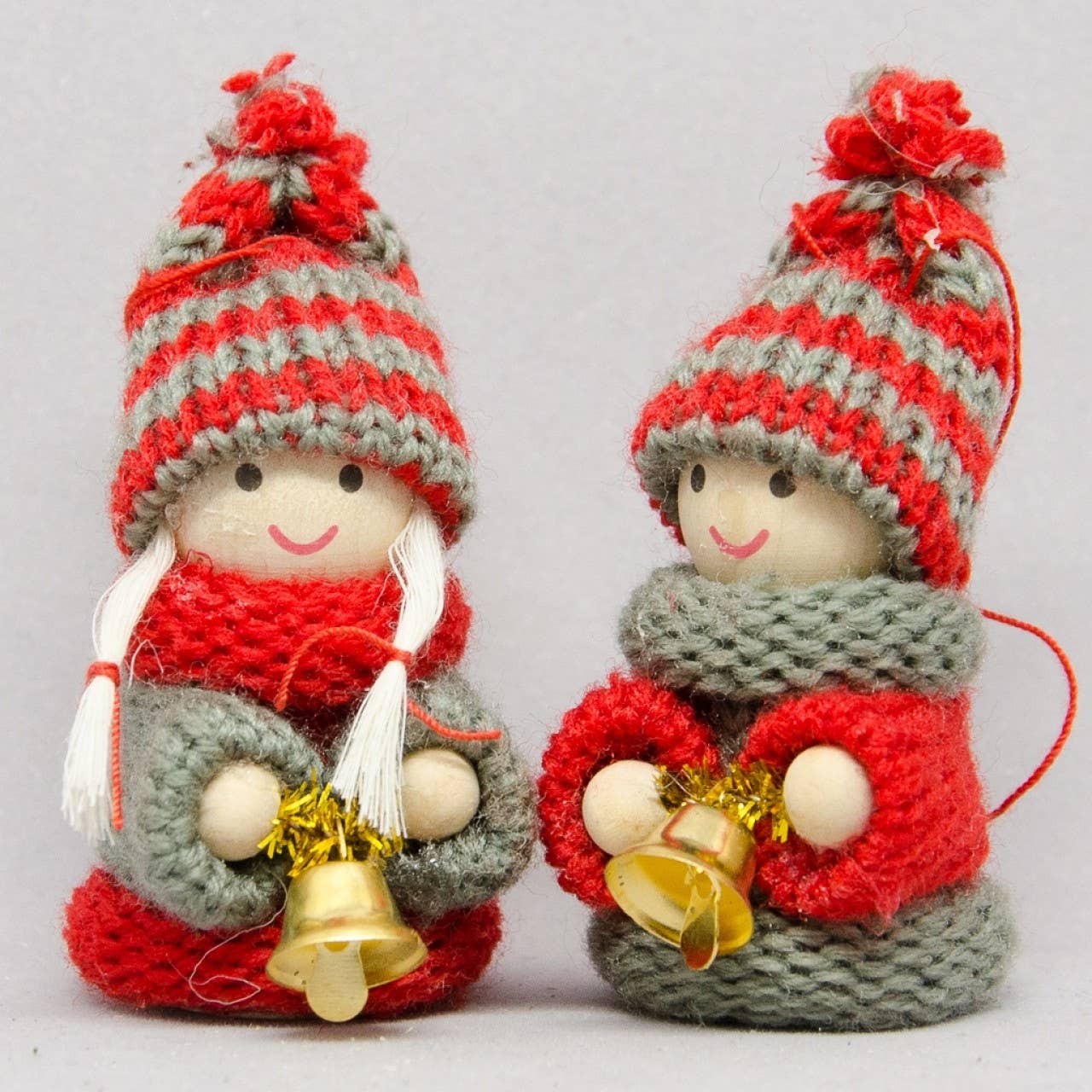 Two Tomte Children with Bells Christmas Tree Ornaments, featuring red and gray knit hats and scarves while holding gold bells, stand side by side—a charming handcrafted wood decor for your festive table.