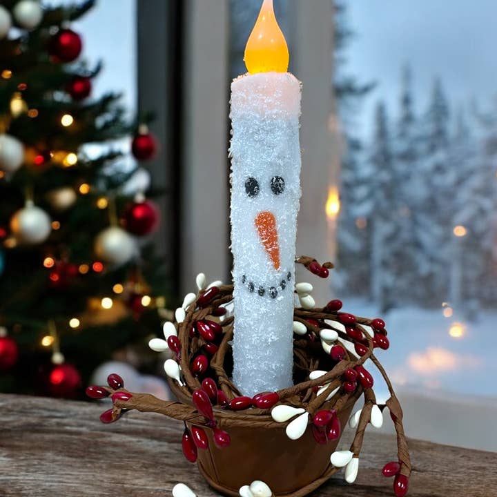 A snowman-shaped candle with a carrot nose and coal buttons is wrapped in a berry garland. Set beside a snowy window and Christmas tree, its illuminated by the warm glow of the Candle: Snowman Face Christmas Battery Operated Timer Taper Candles.