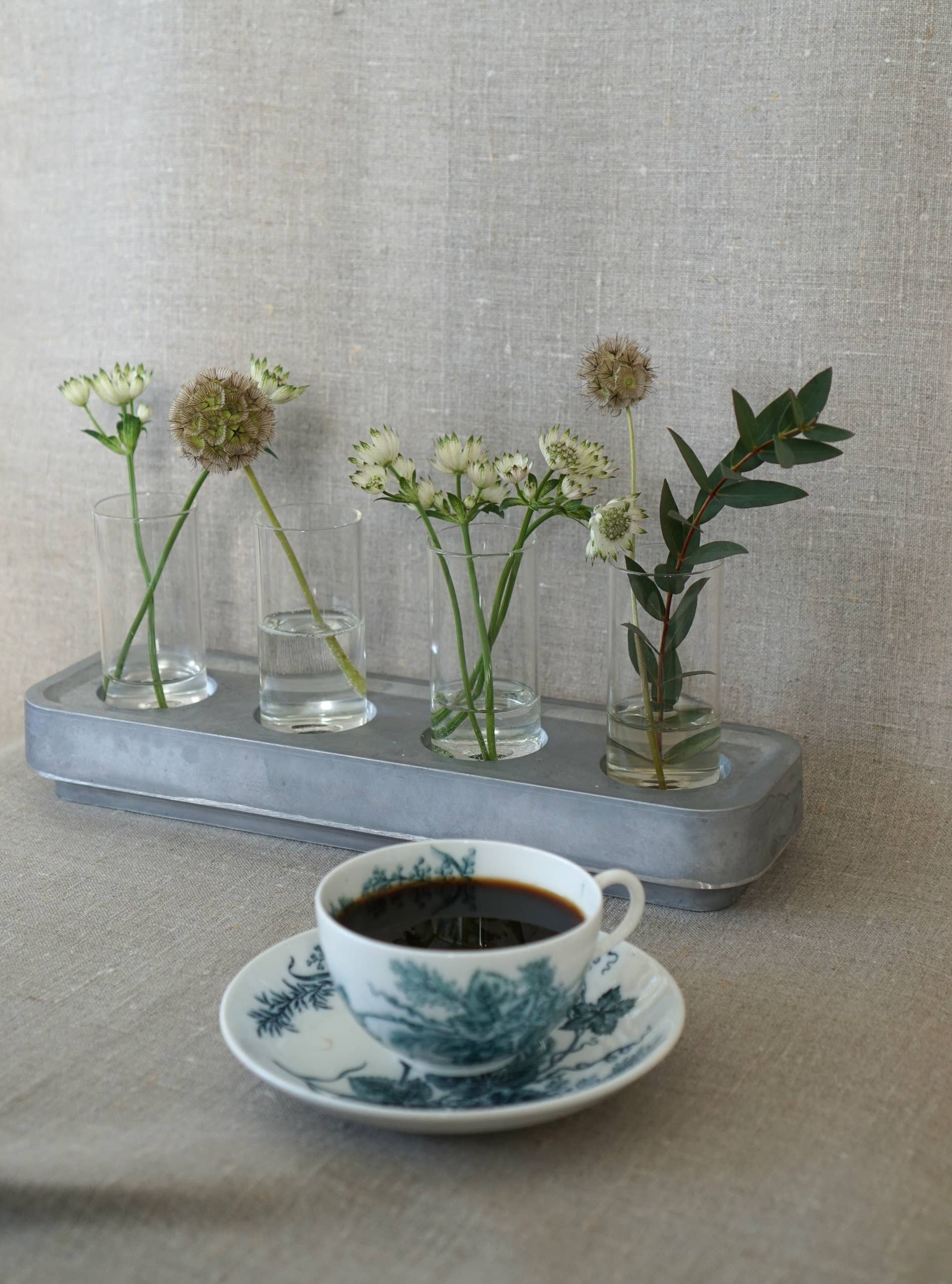 A cup of coffee rests on a blue floral saucer, with four mini vases from the Candle Holder: Mini Vases Pack of 4 for Stumpastaken holding small flowers and greenery. The serene setting is complemented by a textured beige fabric background.