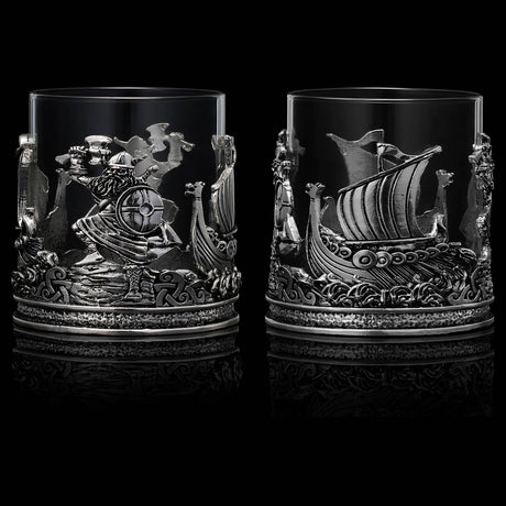 The Viking & Mythological Whiskey Drinking Glass (12 OZ) features two clear glasses with ornate metal bases, showcasing detailed reliefs of ships and warriors against a black background.
