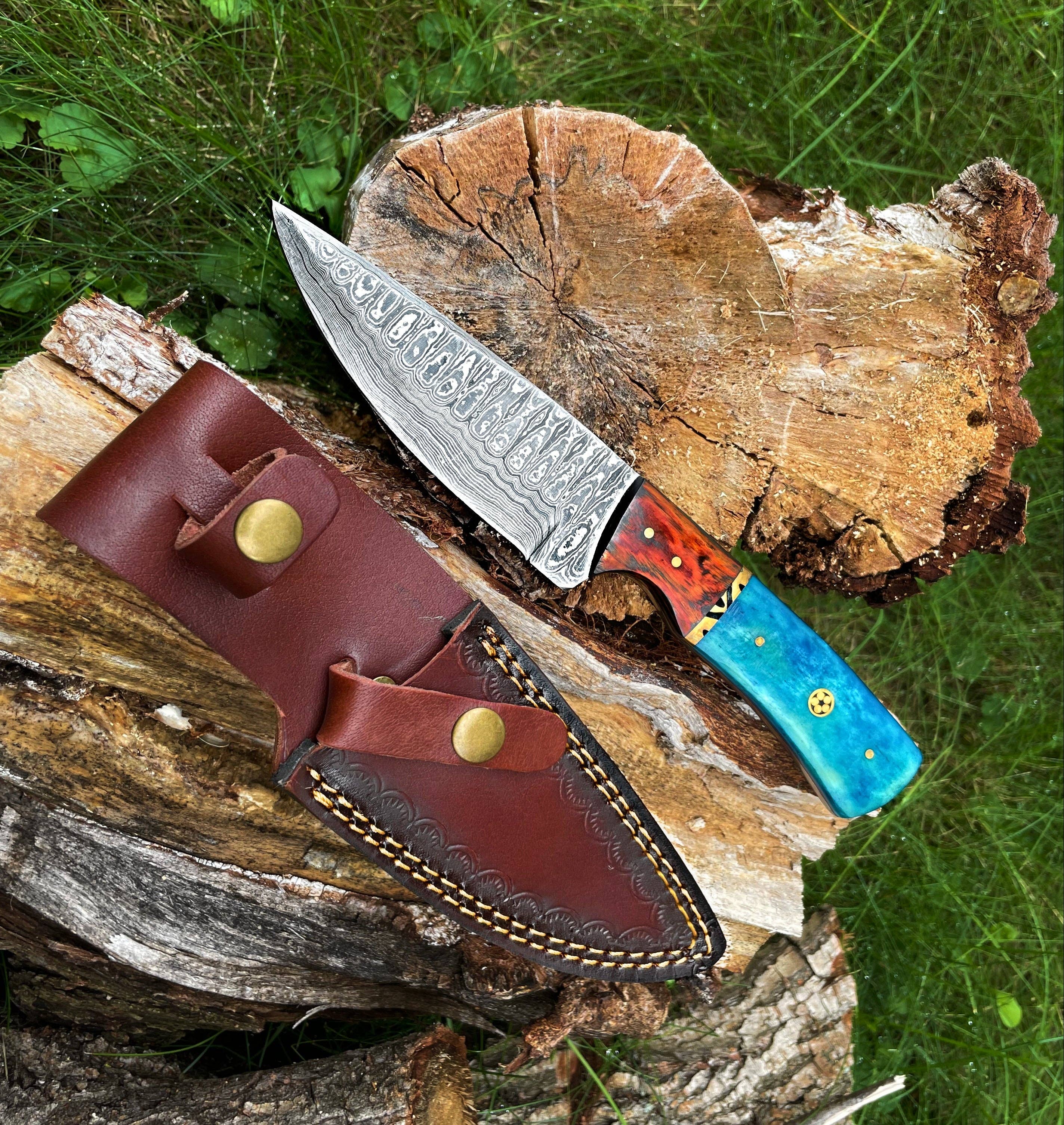 A Hand Forged Damascus Knife with a drop-style blade and black handle rests on a tree stump beside a brown leather sheath, outdoors on grass.
