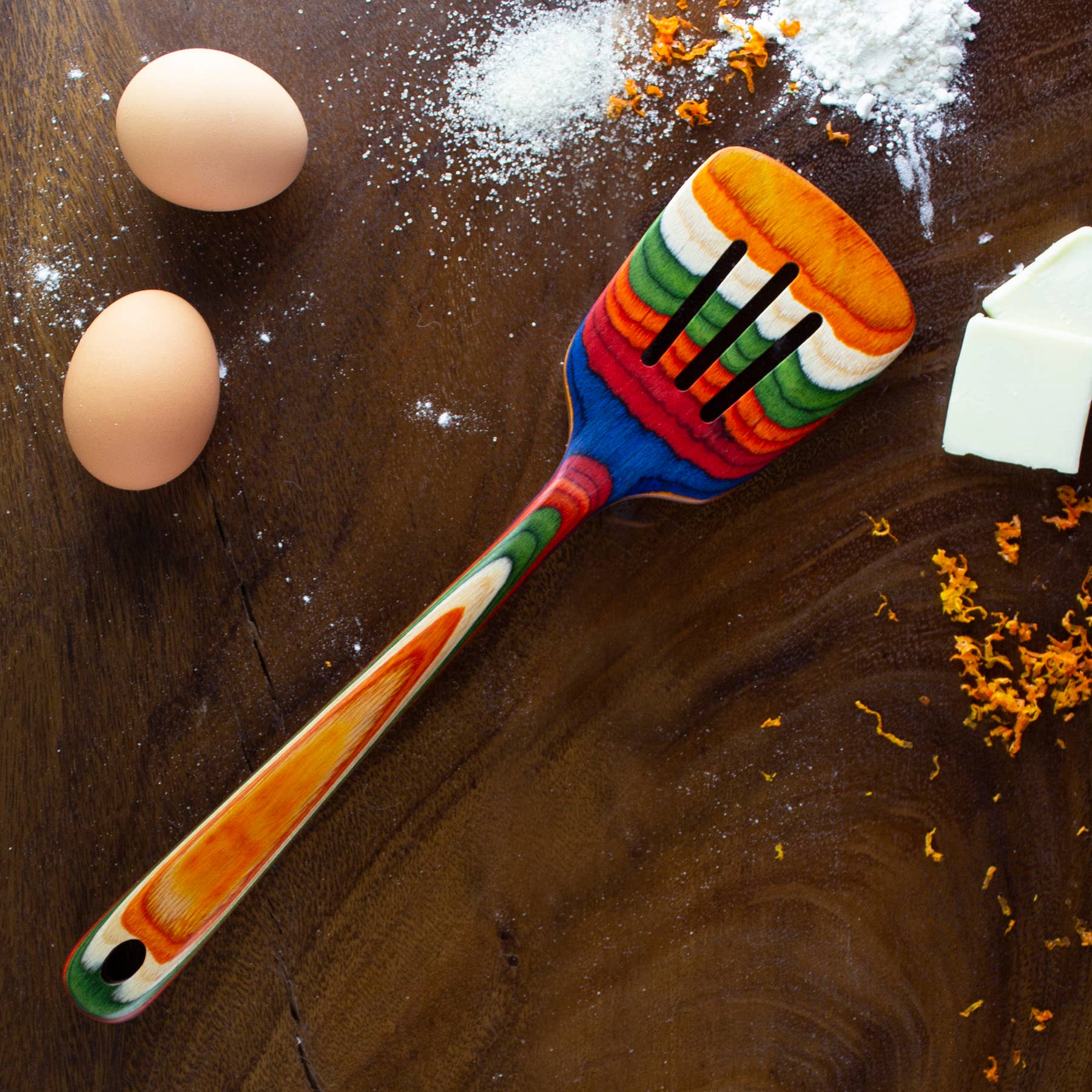 A vibrant wooden slotted spatula from the Marrakesh Collection is placed on a wooden surface, accompanied by two eggs, flour, butter, and orange zest.