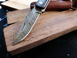 Close-up of the Damascus Steel Pocket knife, featuring a patterned blade and walnut wood handle, resting on a wooden board with its leather sheath in the background.