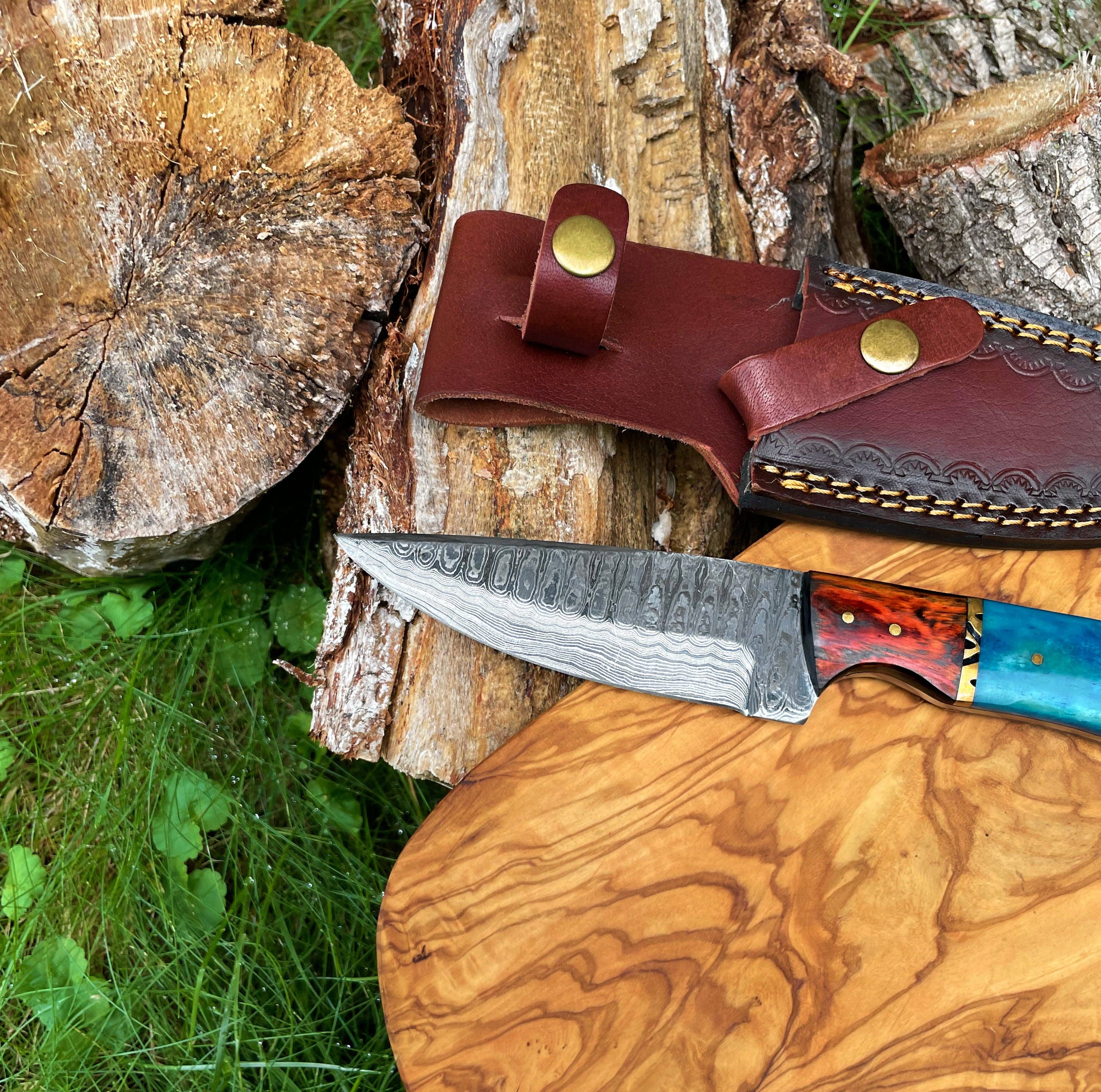 A Hand Forged Damascus Knife with a drop-style blade and black handle lies on a wooden board beside a leather sheath and tree logs on the grass.