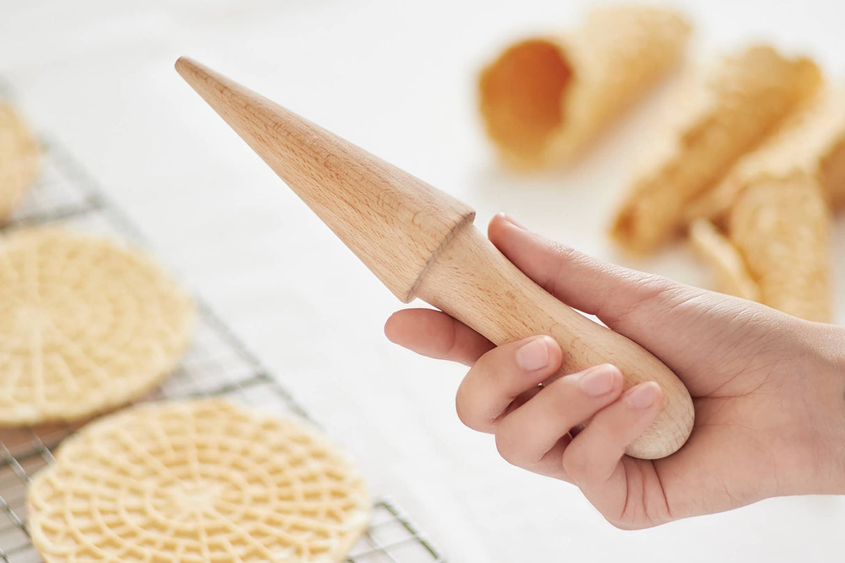 A hand holds the Krumkake Cone Roller (Beechwood, 1.5" x 1.5" x 8.25"), with wafer cookies and empty waffle cones on a cooling rack in the background.