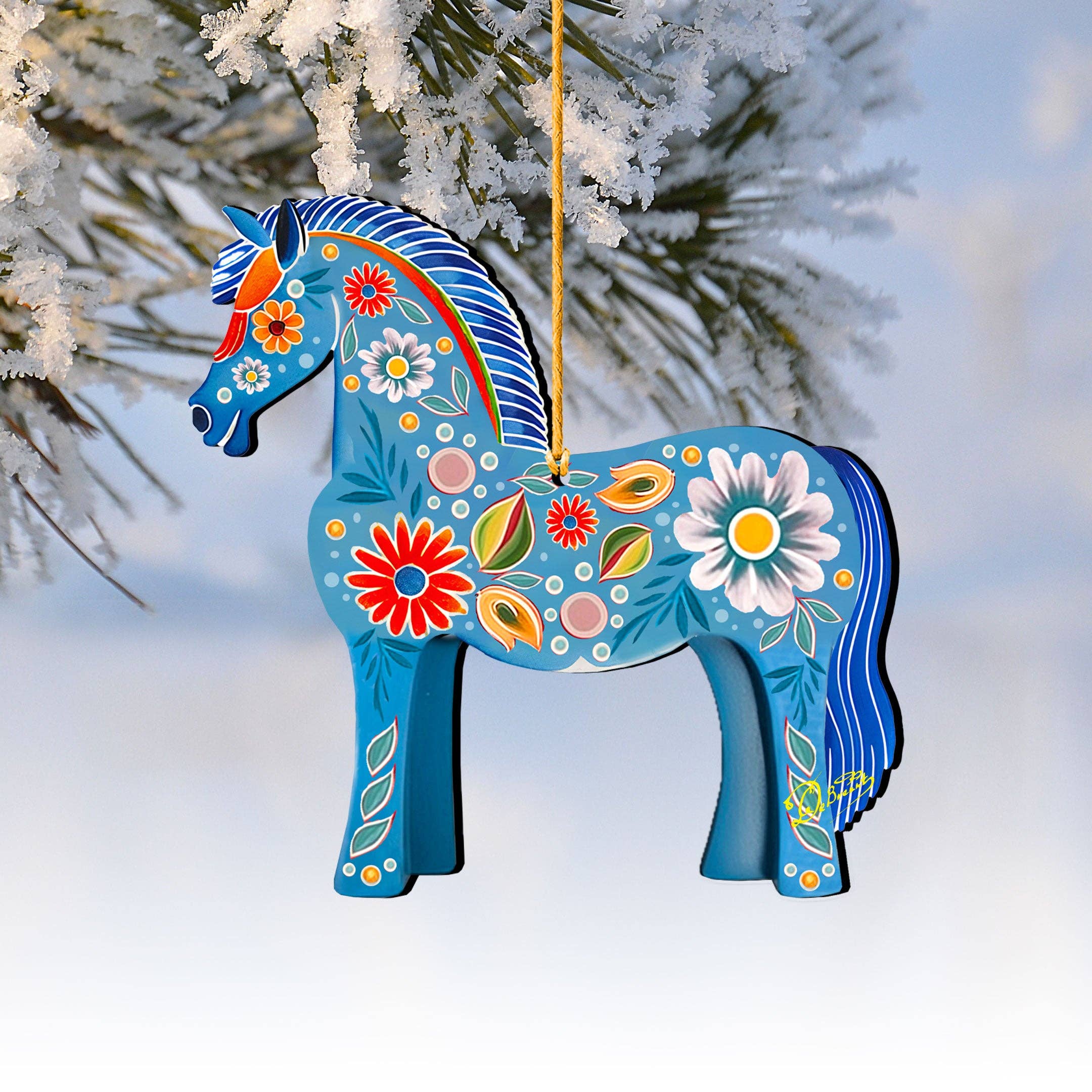 A Danish Horse Decorative Wood Christmas Tree Ornament by G Debrekht (5.5"x5") with colorful floral patterns hangs from a yellow string on a snow-covered branch.