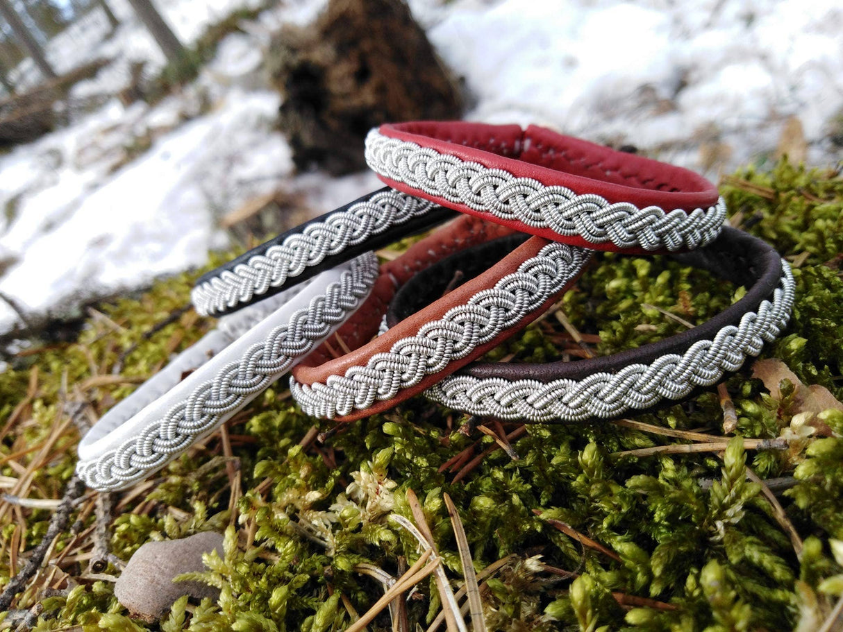Five Traditional Sami Single Braid Bracelets—black, handcrafted in Lapland, Sweden from reindeer leather with metal detailing—rest on mossy ground amid snow and trees.