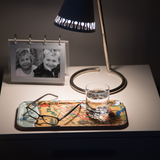 Nightstand with a lamp, birchwood tray holding eyeglasses, framed photo of two children, and a glass of water beside a bed in Scandinavian Hygge style.