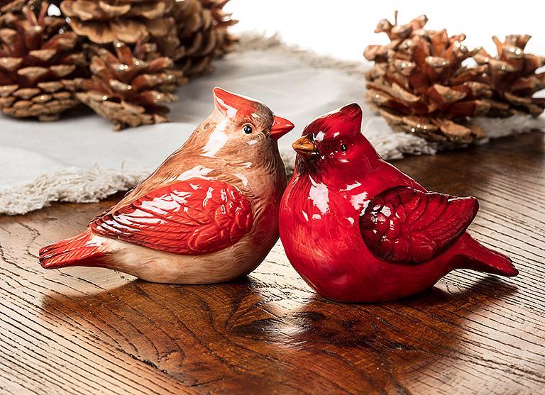 A pair of Shaker: Salt & Pepper Shaker - Cardinals, featuring a vibrant red and a warm brown ceramic bird figurine, is gracefully arranged on a wooden surface, with pinecones adding to the backdrop's charm.