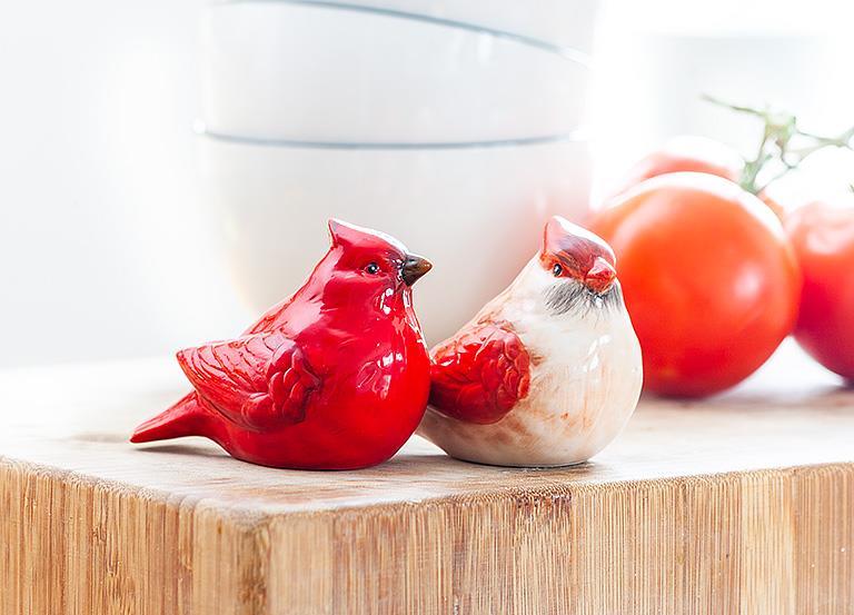 A set of vibrant cardinal-themed ceramic salt and pepper shakers, featuring a red cardinal and white shaker with red accents, is elegantly displayed on a wooden surface. In the softly focused background, bowls and tomatoes enhance the scene's vivid charm.