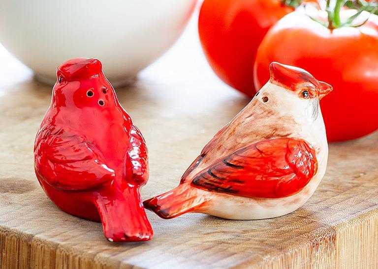 A pair of "Shaker: Salt & Pepper Shaker - Cardinals" rests on a wooden surface, their vibrant red and orange tones echoing the colorful ripe tomatoes in the background.