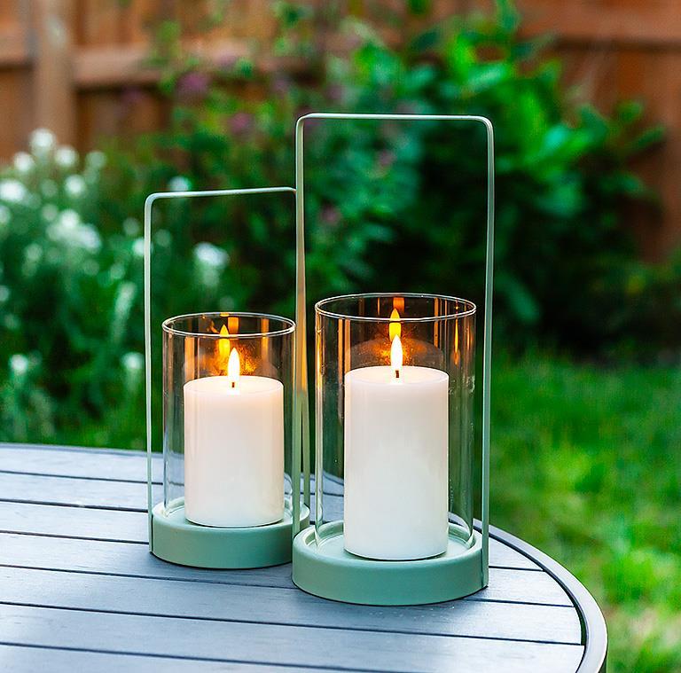 Two lit candles in 10.5" cylinder lanterns adorn a round outdoor table, enhancing the garden's charm with stylish outdoor decor.