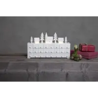 The Candelabra: Star Trading Yuletide Advent Calendar, featuring a snow-covered village scene, sits on a gray surface. Its tiny doors invite curiosity as LED lights gently illuminate the wrapped gifts and nearby small plant.