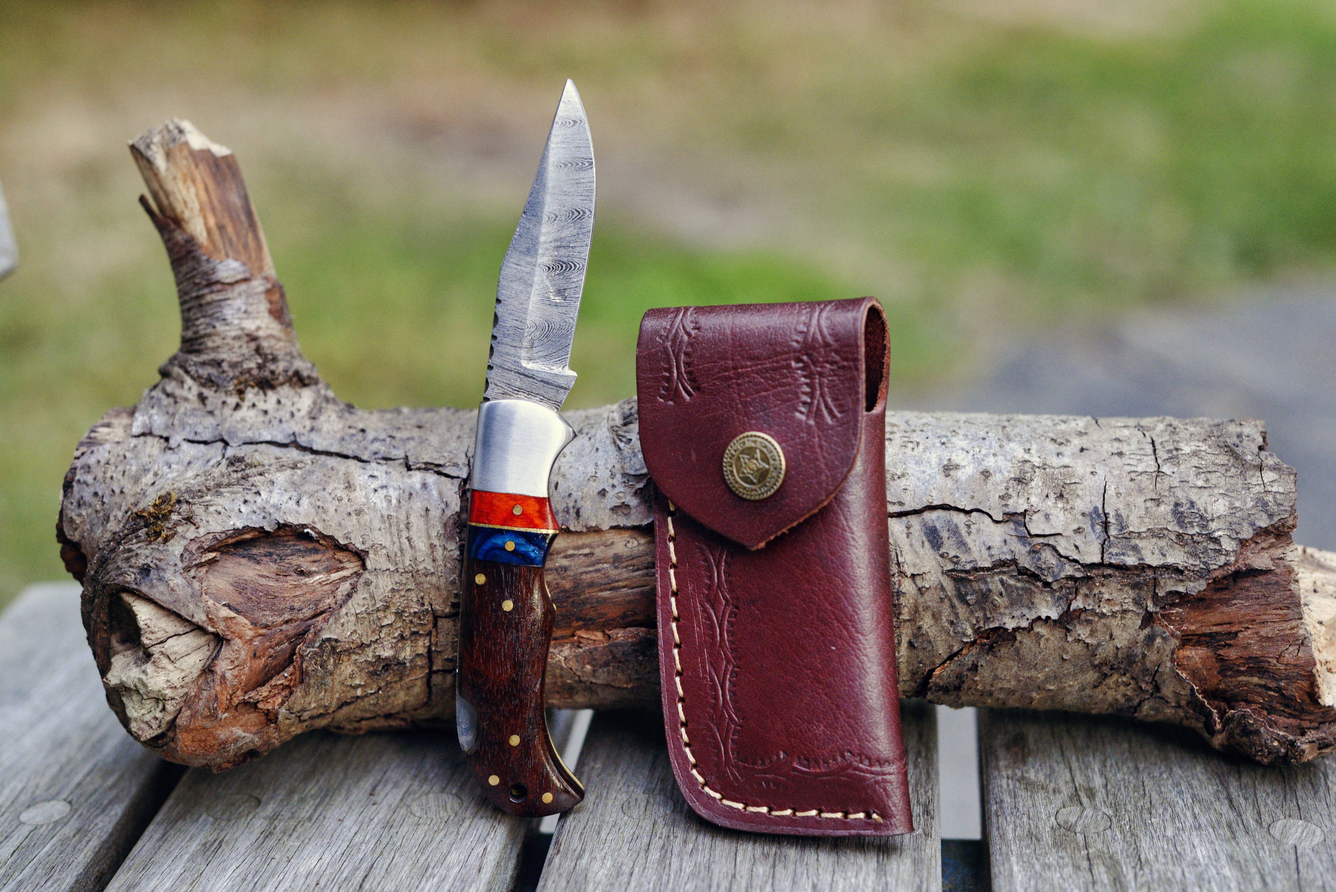 A Red/Black Damascus folding pocket knife with a patterned blade and a wooden handle is elegantly displayed on a piece of wood, accompanied by a brown leather sheath.