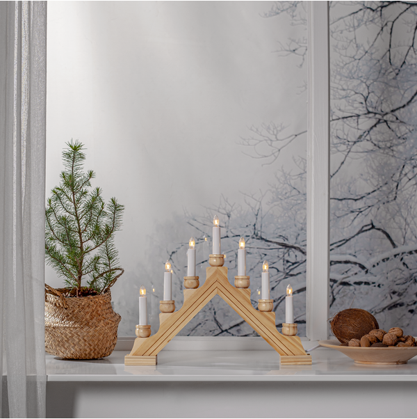 A small pine tree in a woven basket sits beside a Star Trading - Karin Electric 7-Light candelabra, casting a warm glow against the snowy landscape outside. Nuts and shells are arranged on the right, completing this cozy scene.