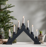 The Candelabra: Star Trading - Karin Electric 7-Light sits on a white surface, its black candle arch echoing traditional Swedish Christmas candlesticks. In the background, a small evergreen tree and potted bulb plant add charm.