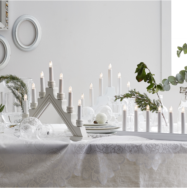 A table is set with the electric 7-light Candelabra from Star Trading's Karin collection, accompanied by a lace tablecloth and clear glass ornaments, embodying Swedish Christmas tradition. Plates and green foliage enhance the indoor decoration against a white wall backdrop.