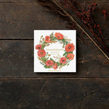 A square card folder sits on wood, featuring a floral wreath of red poppies and green leaves. Named Card Pack: Summer Wreaths by Koustrup & Co - Denmark, it is elegantly adorned with a branch of red berries for added charm.