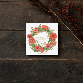 A square card folder sits on wood, featuring a floral wreath of red poppies and green leaves. Named Card Pack: Summer Wreaths by Koustrup & Co - Denmark, it is elegantly adorned with a branch of red berries for added charm.