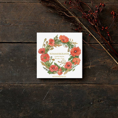 A square card folder sits on wood, featuring a floral wreath of red poppies and green leaves. Named Card Pack: Summer Wreaths by Koustrup & Co - Denmark, it is elegantly adorned with a branch of red berries for added charm.