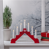 The Star Trading Adam 7 Bright Red Floral Candelabra with seven glowing candles sits on a white windowsill beside a potted evergreen and wrapped gifts, creating charming window decor against the snowy backdrop.