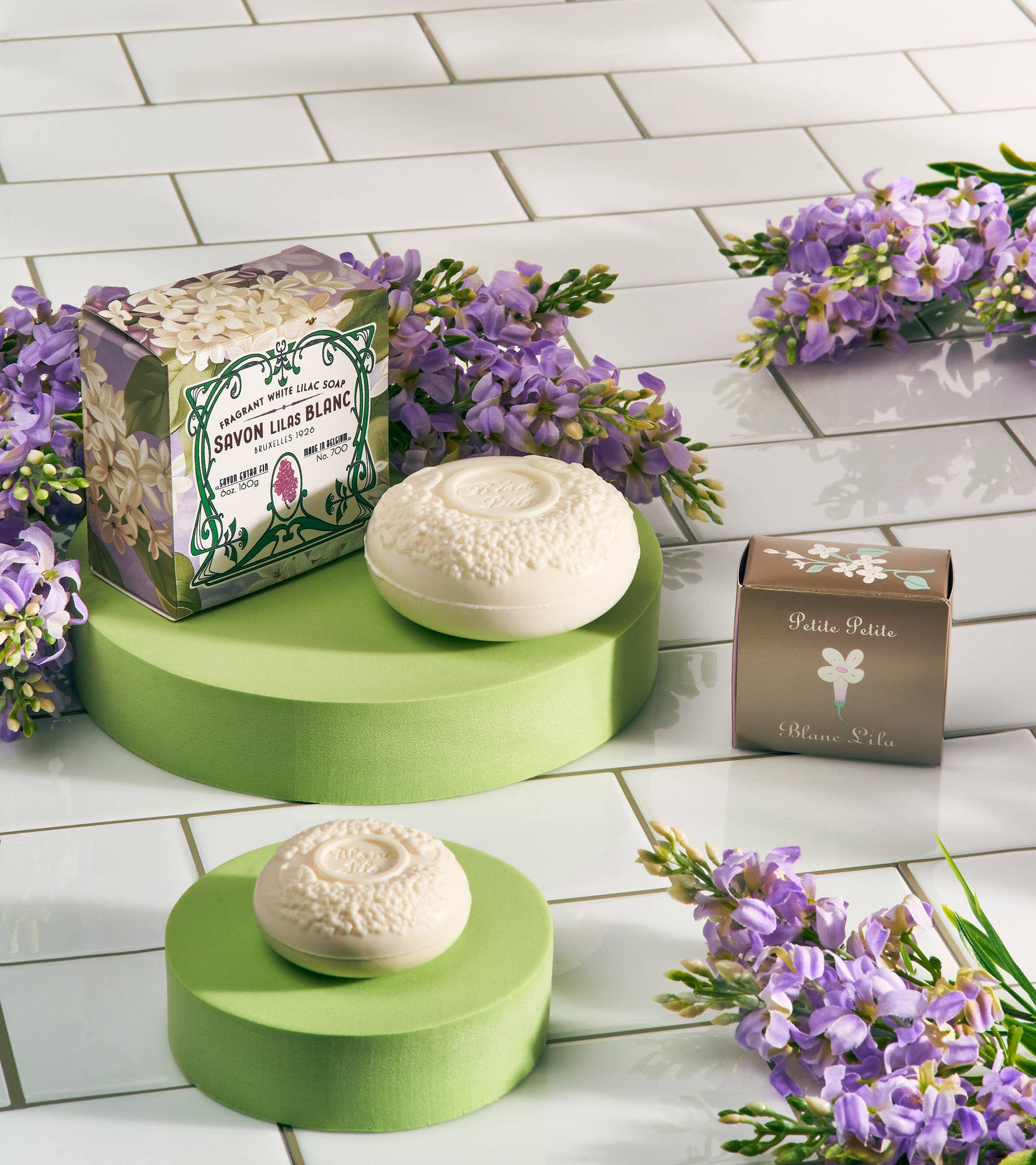 Embossed soap bars are showcased on green platforms. One is a Savon Lilas Blanc Fragrant White Lilac Bar Soap in a floral box, with purple flowers scattered on white tiles. This vegan collection adds elegance to any setting with its delicate touch.