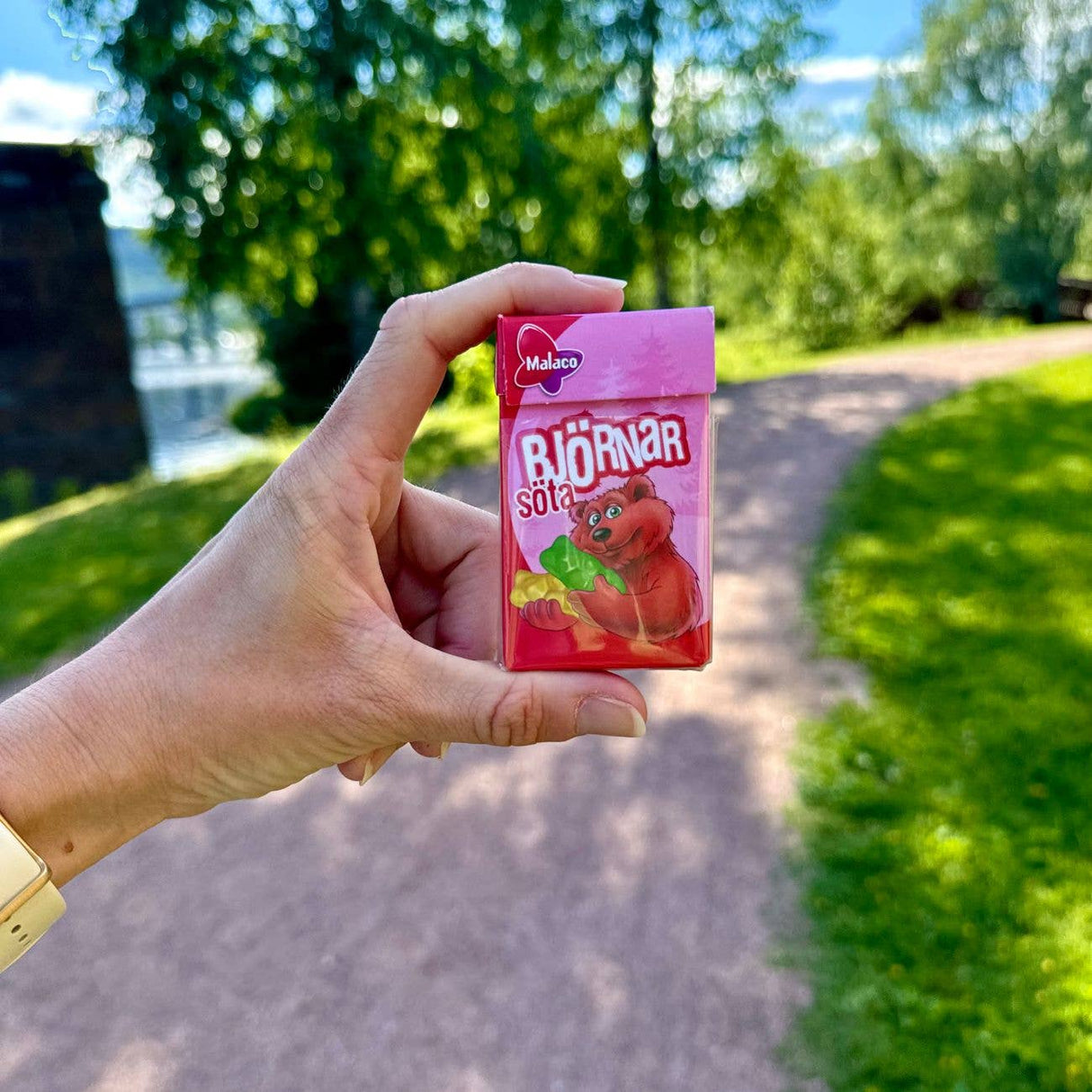 A hand holding a pink 32g mini box of Malaco Bjornar Sota Sweet Bears by Swedish Sweets Wholesale LLC - Swedish Candy outdoors, with a sunny park path and green trees in the background.