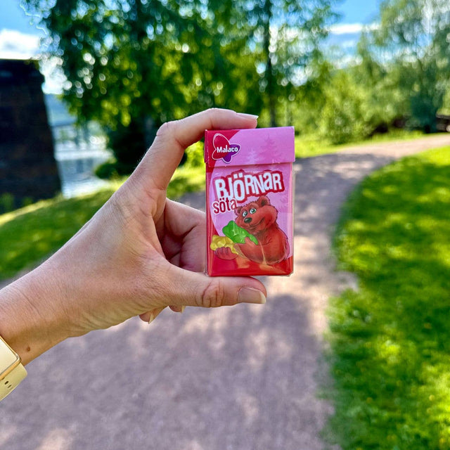 A hand holding a pink 32g mini box of Malaco Bjornar Sota Sweet Bears by Swedish Sweets Wholesale LLC - Swedish Candy outdoors, with a sunny park path and green trees in the background.