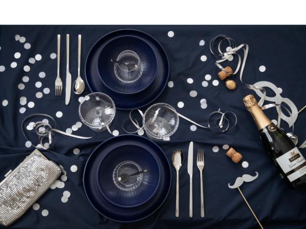 A festive table setting includes two blue plates, two glasses, a sparkling wine bottle, and the Gense: Still Cutlery Set in Matte/Polished Steel. Adorned with confetti, silver ribbons, a sparkling clutch, and mustache props on a dark tablecloth, it exudes Nordic design charm.