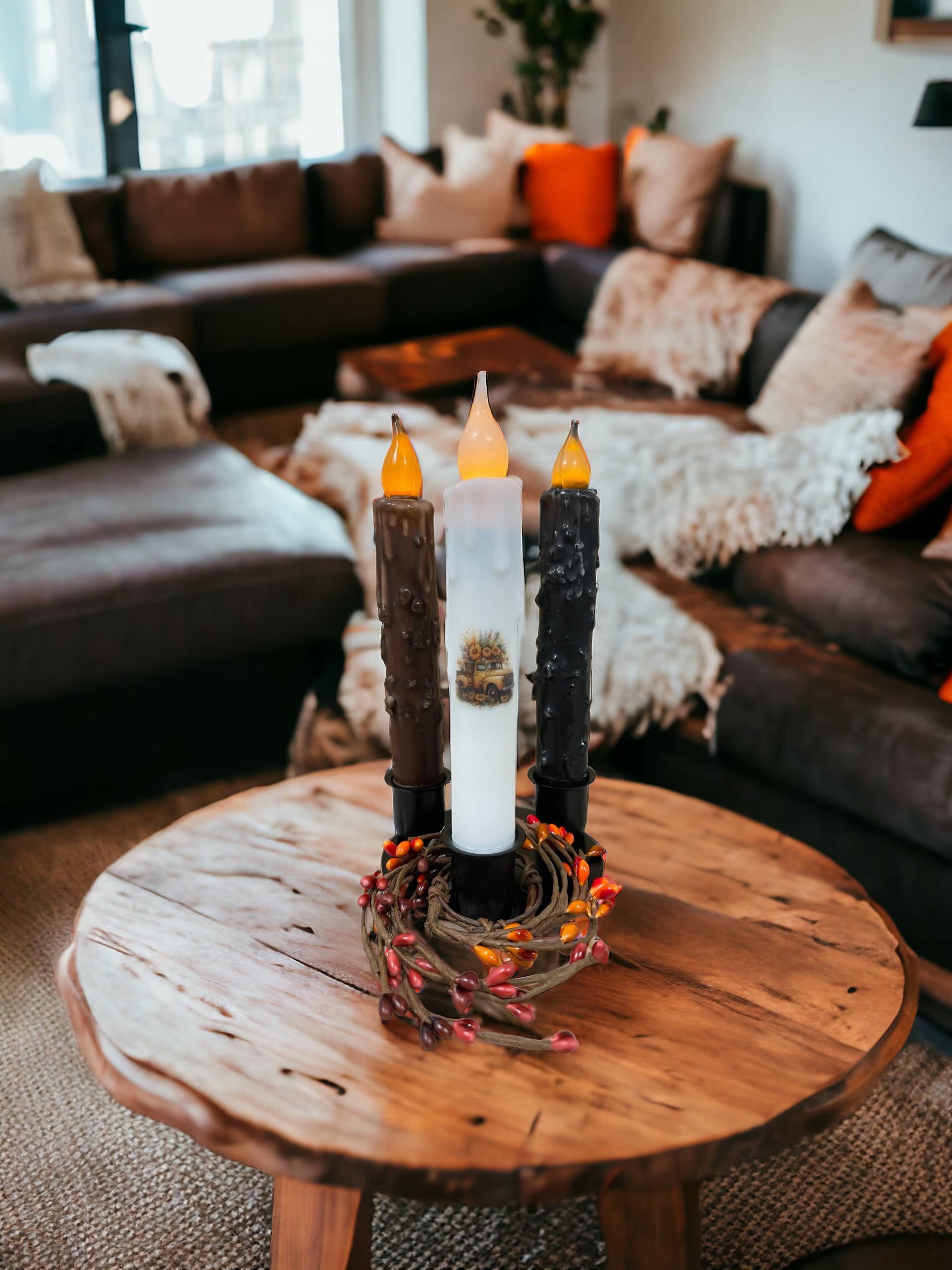 Three LED candles rest on a wooden table, nestled in a decorative wreath and held by a Round Triple Wrought Iron Taper Holder. The cozy living room, with its inviting sofas and blankets, showcases the craftsmanship of Lancaster blacksmith products.