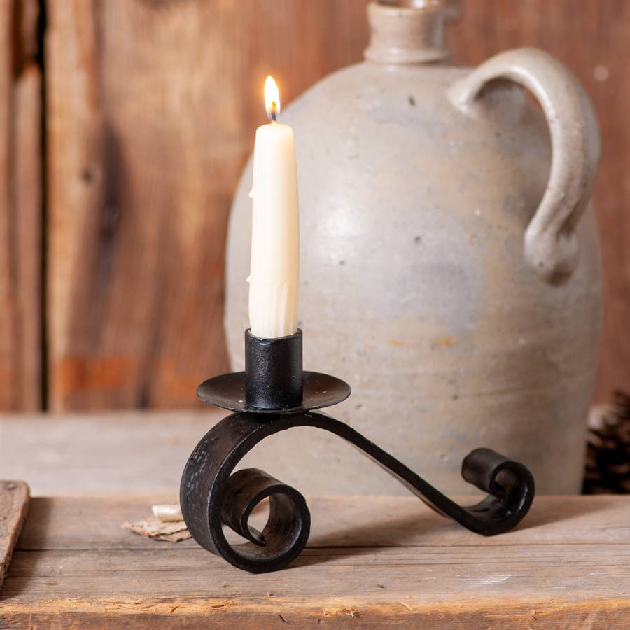 A lit white candle rests in the Wrought Iron Curled Pillar Candle Holder on a wooden surface, with a ceramic jug and rustic wood background behind it.