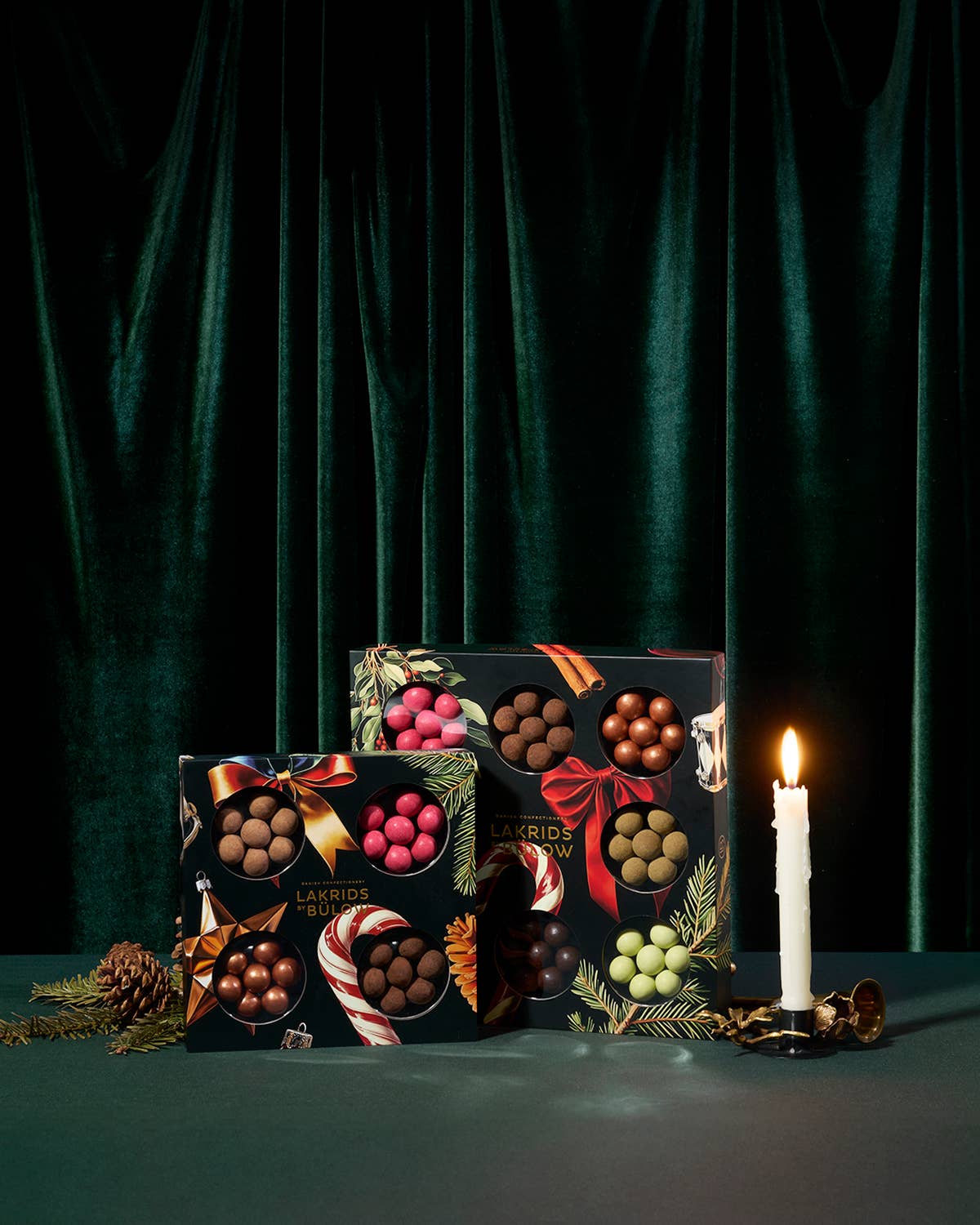 Two Lakrids by Bulow Winter Collection 4 Flavor Selection Boxes (12.3 oz each) are arranged beside a glowing white candle, pinecones, and festive greenery on a dark green velvet backdrop.