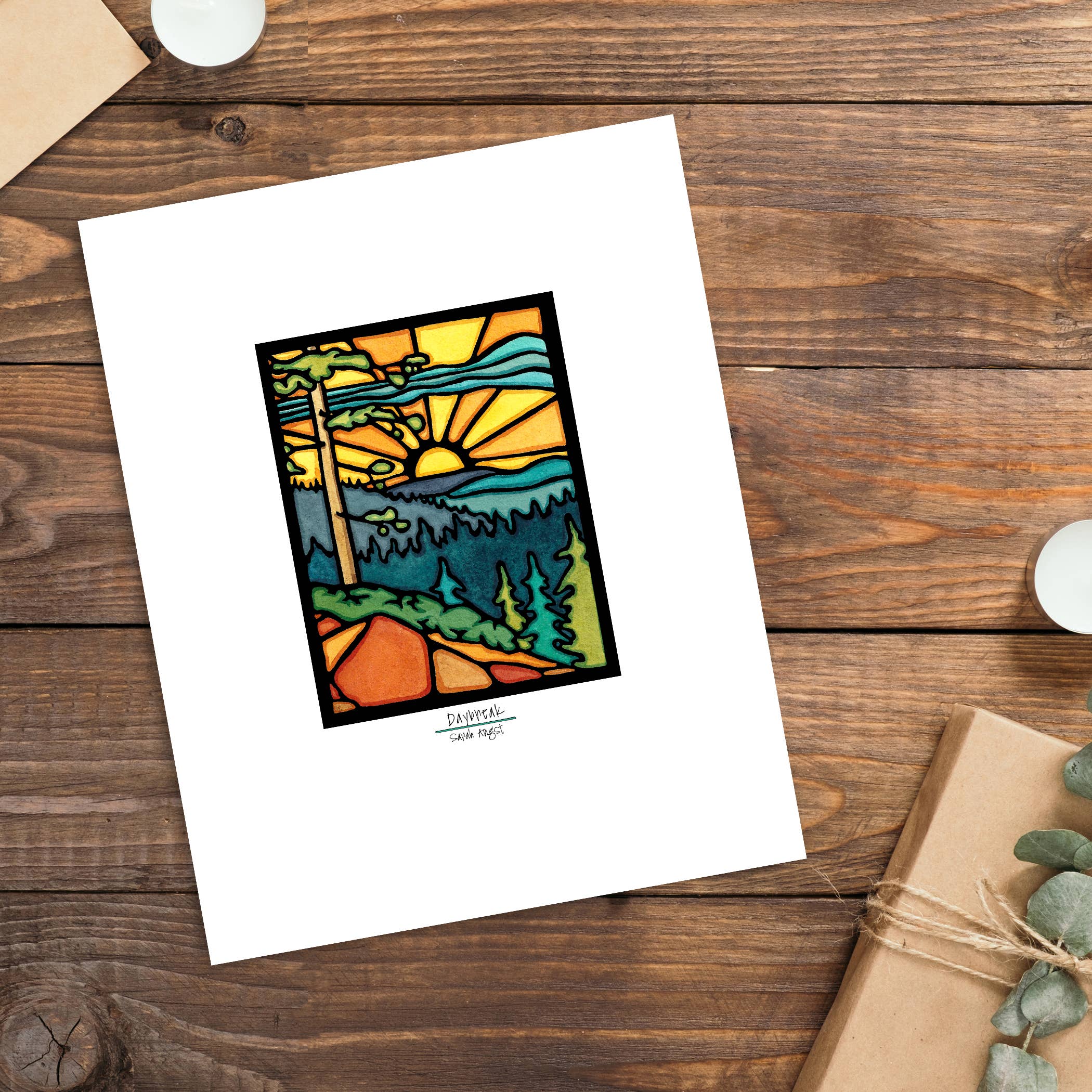 Artwork: "Daybreak: 11"x14" by Sarah Angst," a stained glass depiction of a sunlit landscape featuring a winding path, trees, and a vibrant sunrise, is available as Giclee Reproduction. It is elegantly displayed on a wooden table with a candle and wrapped gift beside it.