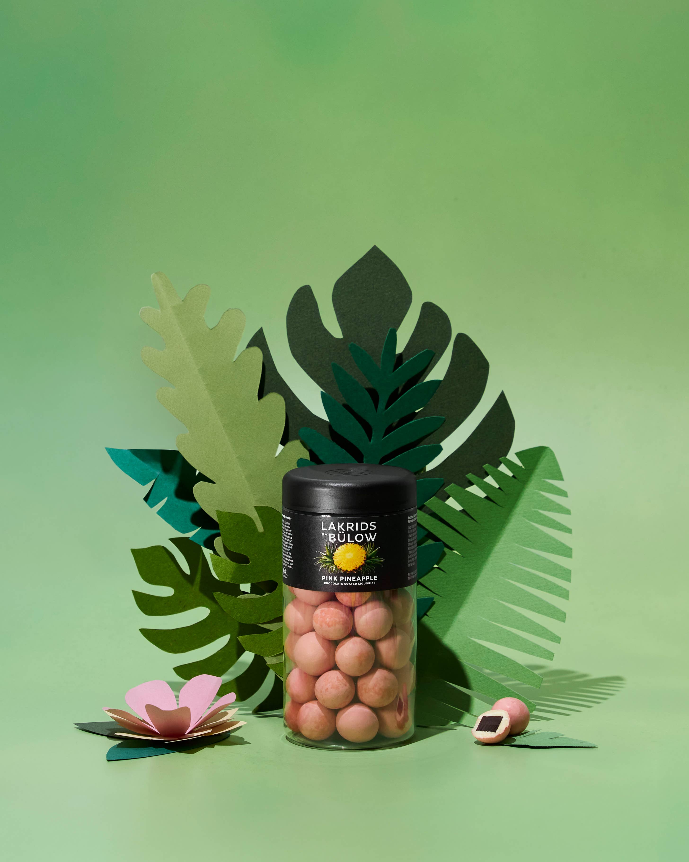 A jar of Lakrids by Bülow - Pink Pineapple Danish Chocolate Coated Licorice (125g) sits in front of paper-cut green leaves, with one candy and a flower beside it—a vibrant treat for licorice and chocolate lovers.