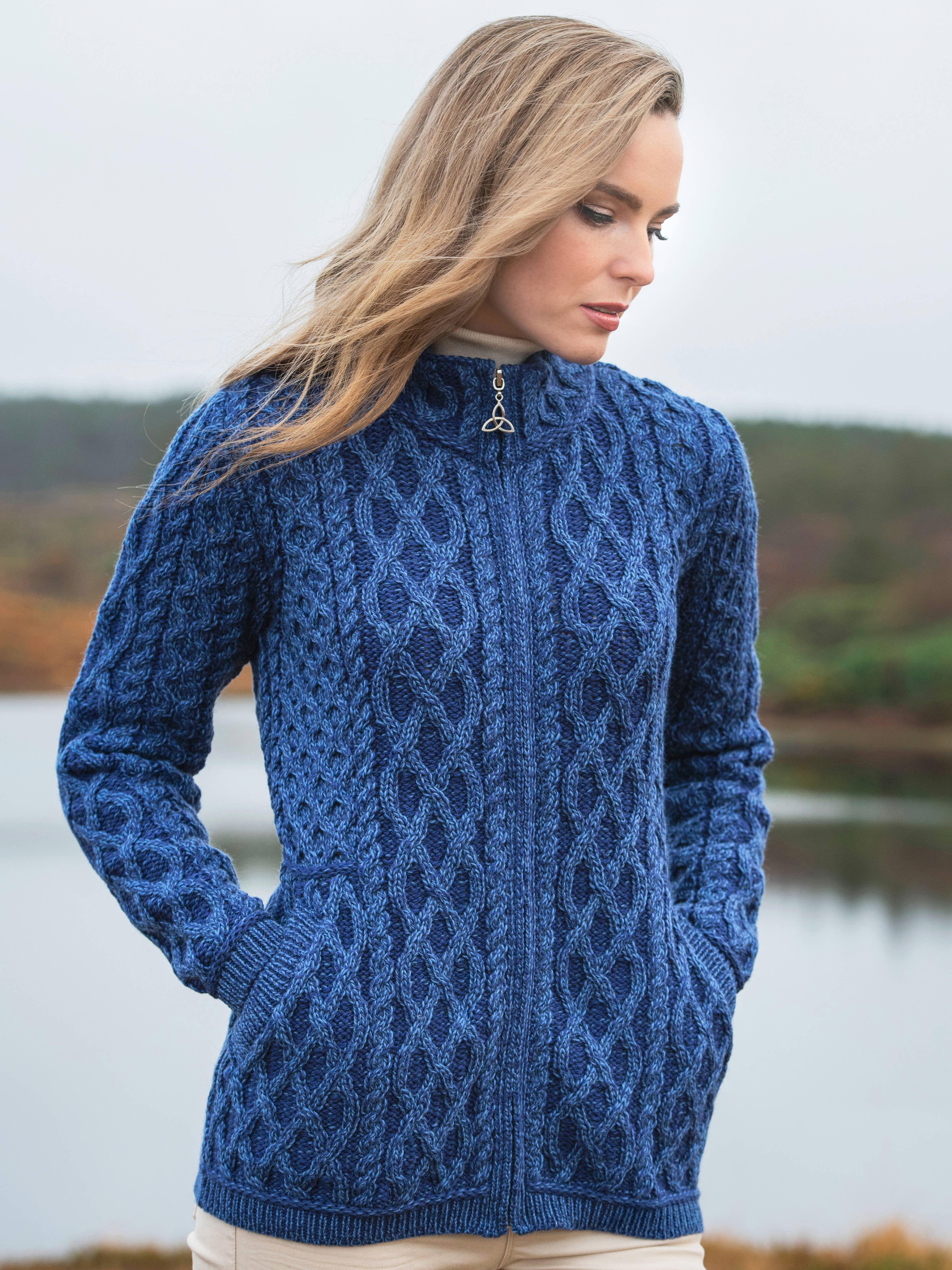 A woman stands outdoors near water, wearing the Plated Zip Cardigan - Marl Blue Mix sweater with her hands in the pockets, looking down and to the side.