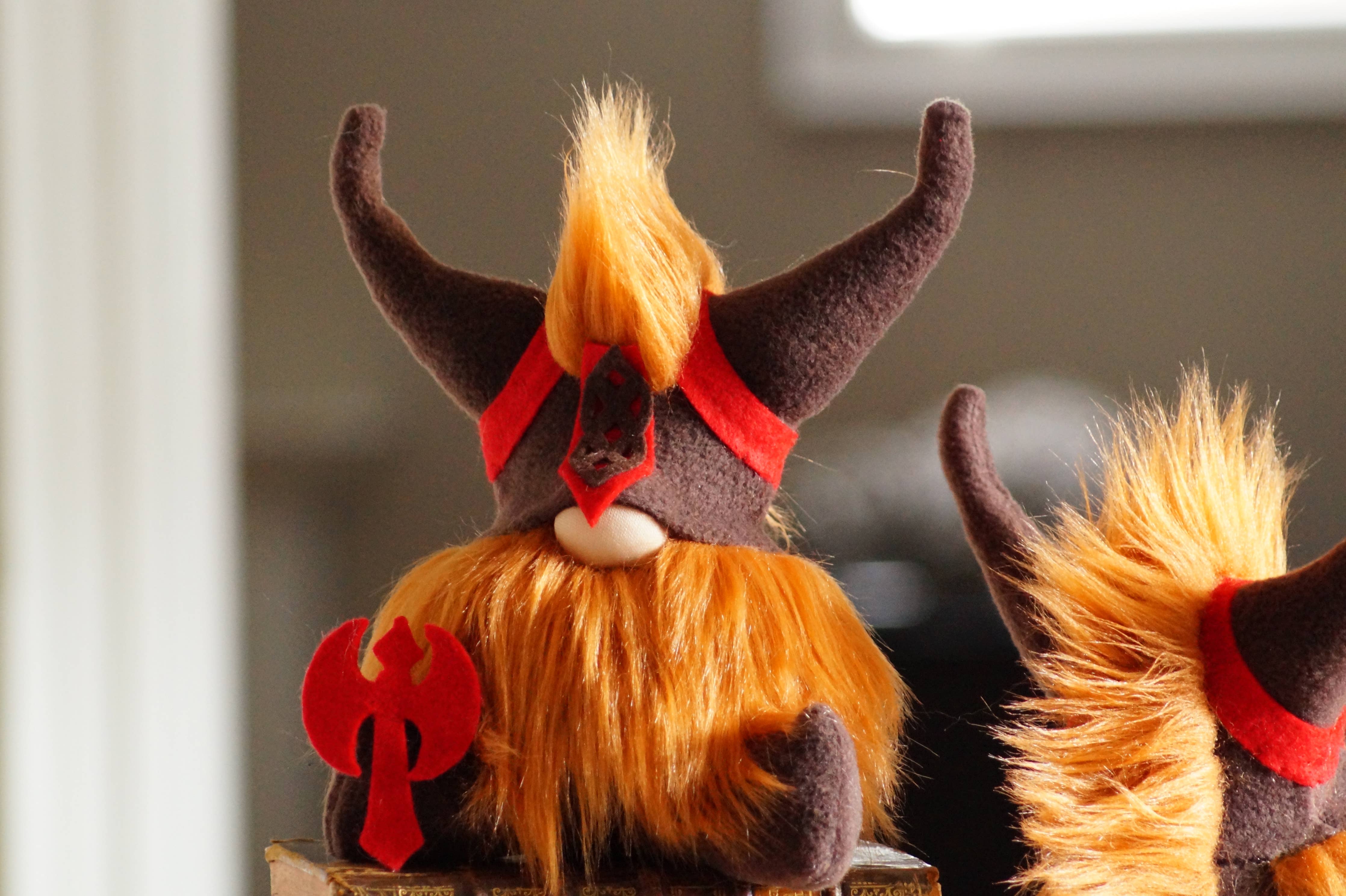 Displayed indoors is a plush creature known as the Gnome: Viking Gnome, featuring orange fur, black and red horns, and holding a small red axe—a unique gift for fantasy lovers.