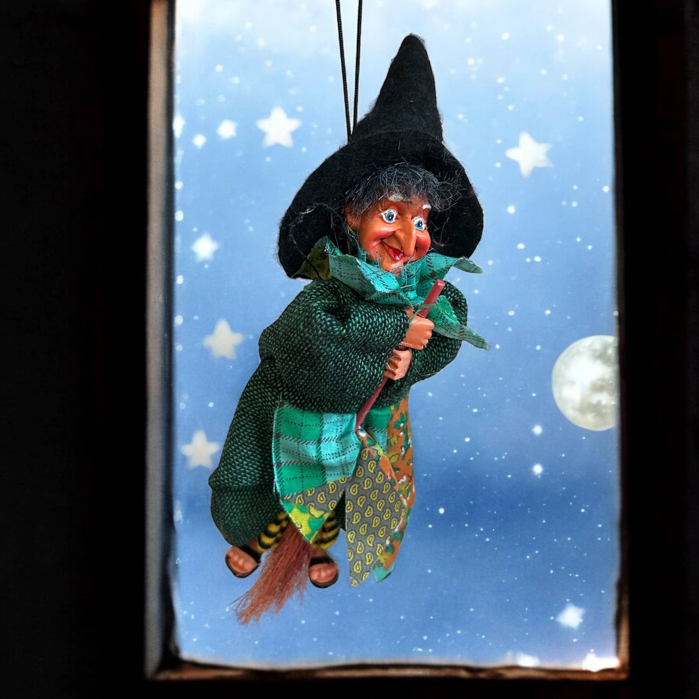 The Johanna Green Kitchen Witch Doll, dressed in green with a black hat, hangs by a string before a full moon and starry sky. Handmade in Germany, this kitchen witch is a charming good luck symbol for your home.