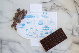 Chocolate bar and cocoa beans on marble surface next to illustrated packaging with a “OUR JOURNEY” map, featuring Scandinavian design elements.