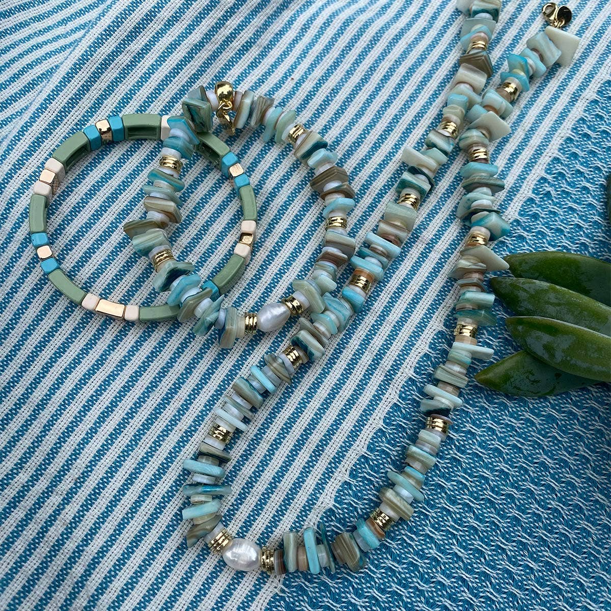 Explore the Wild Nature Collection with the A&C Oslo Blue Shell Necklace, a stunning turquoise beaded piece accented with gold and pearls. It is beautifully presented on textured, blue-striped fabric that evokes the essence of semi-precious stones.