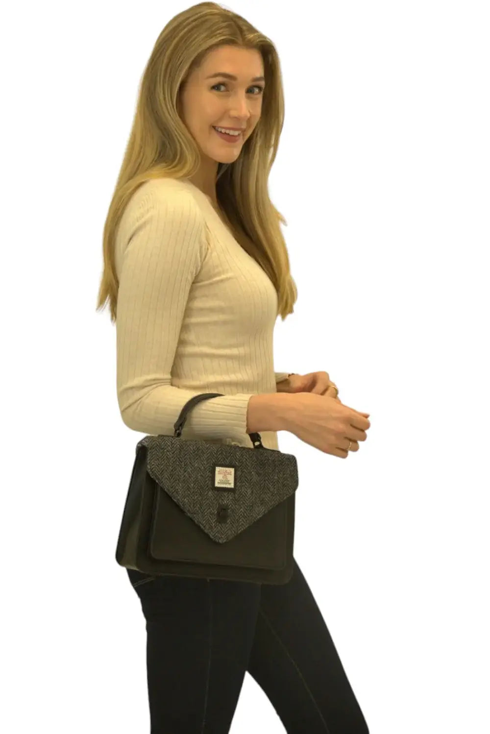 A woman with long blonde hair wears a cream ribbed sweater and dark pants, holding the Harris Tweed Large Satchel - Grey, featuring a textured gray flap crafted from handwoven Harris Tweed.
