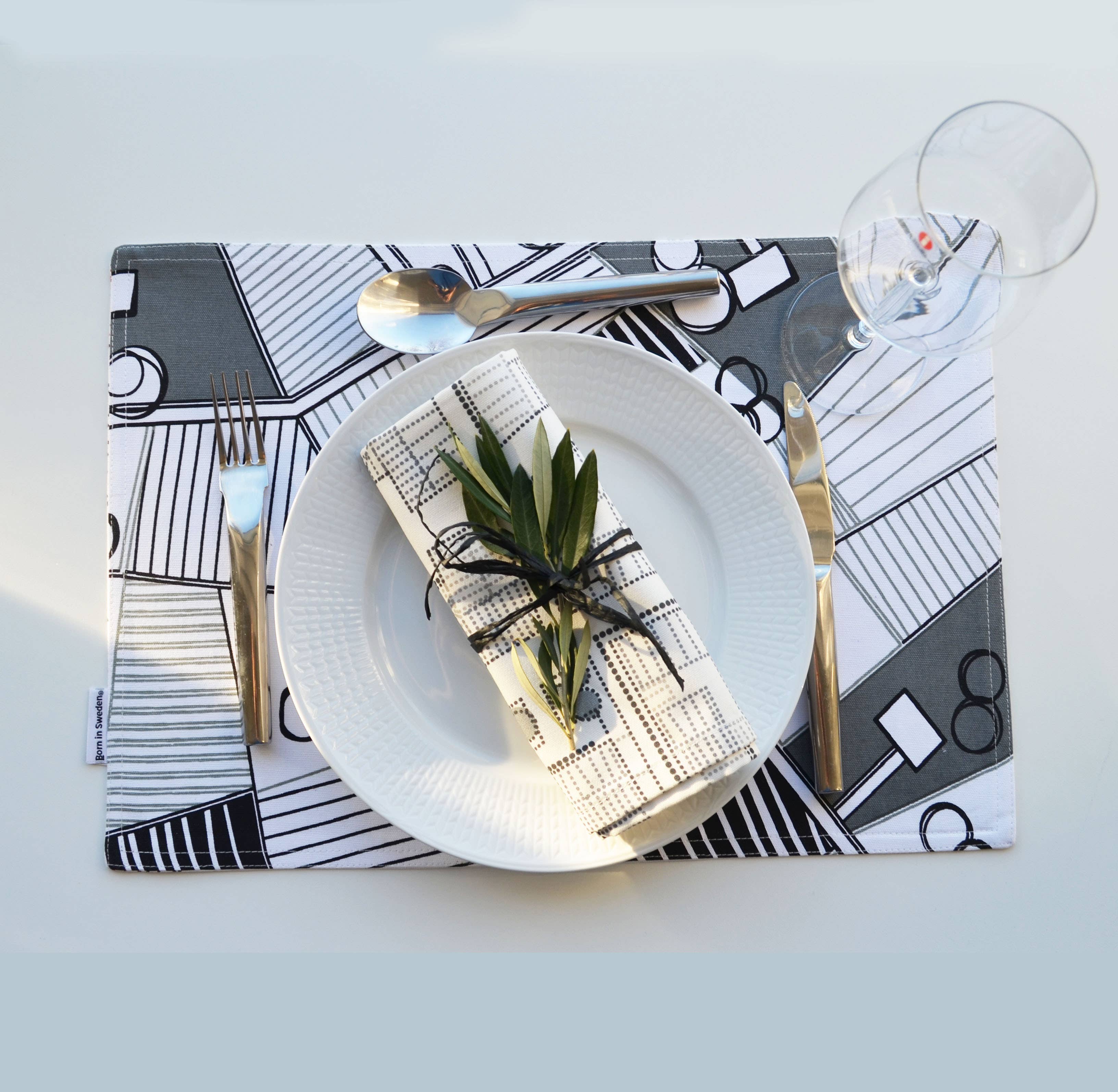 A table setting showcases a white plate with a black and white patterned napkin tied with twine and a sprig, placed on Born in Sweden's double-sided geometric placemats, measuring 47cm x 34cm. Nearby, a fork, knife, spoon, and an empty glass are arranged.