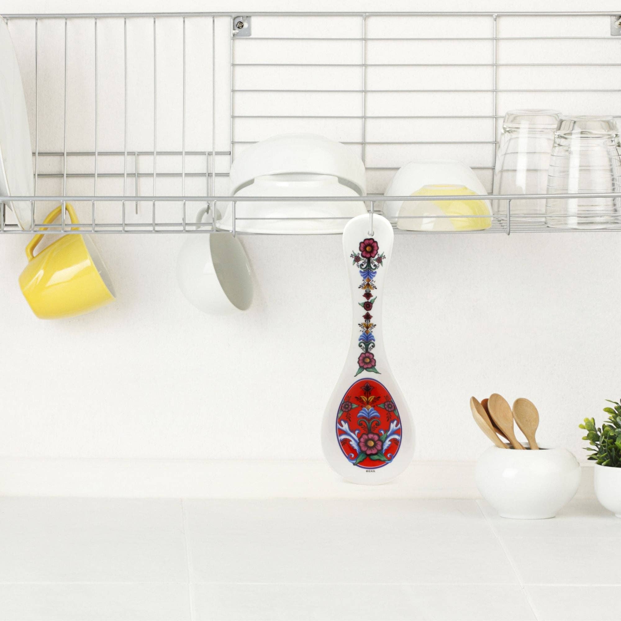 In a kitchen scene exuding Norwegian elegance, a Spoon Rest: Rosemaling Red Kitchen is displayed hanging from a rack, while cups and glasses line the shelf and wooden spoons rest in a bowl on the counter.