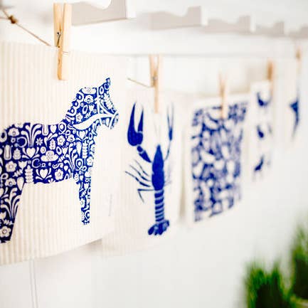 Product Name: Dish Cloth: Dala Horse Blue 
Description: The Dish Cloth in Dala Horse Blue features charming animal designs, such as a horse and a lobster, hanging on a line with clothespins. Made from biodegradable materials, these cloths provide an eco-friendly solution for your everyday cleaning tasks.