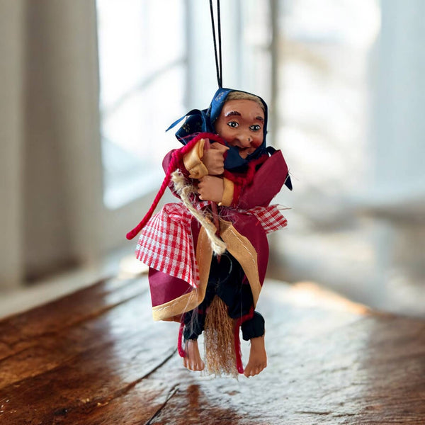 The Paolina Red Kitchen Witch Doll – Broom Rider (6" Tall) is a handmade German figurine in a red cloak and checkered apron, hanging indoors by a string as a charming kitchen protection charm.