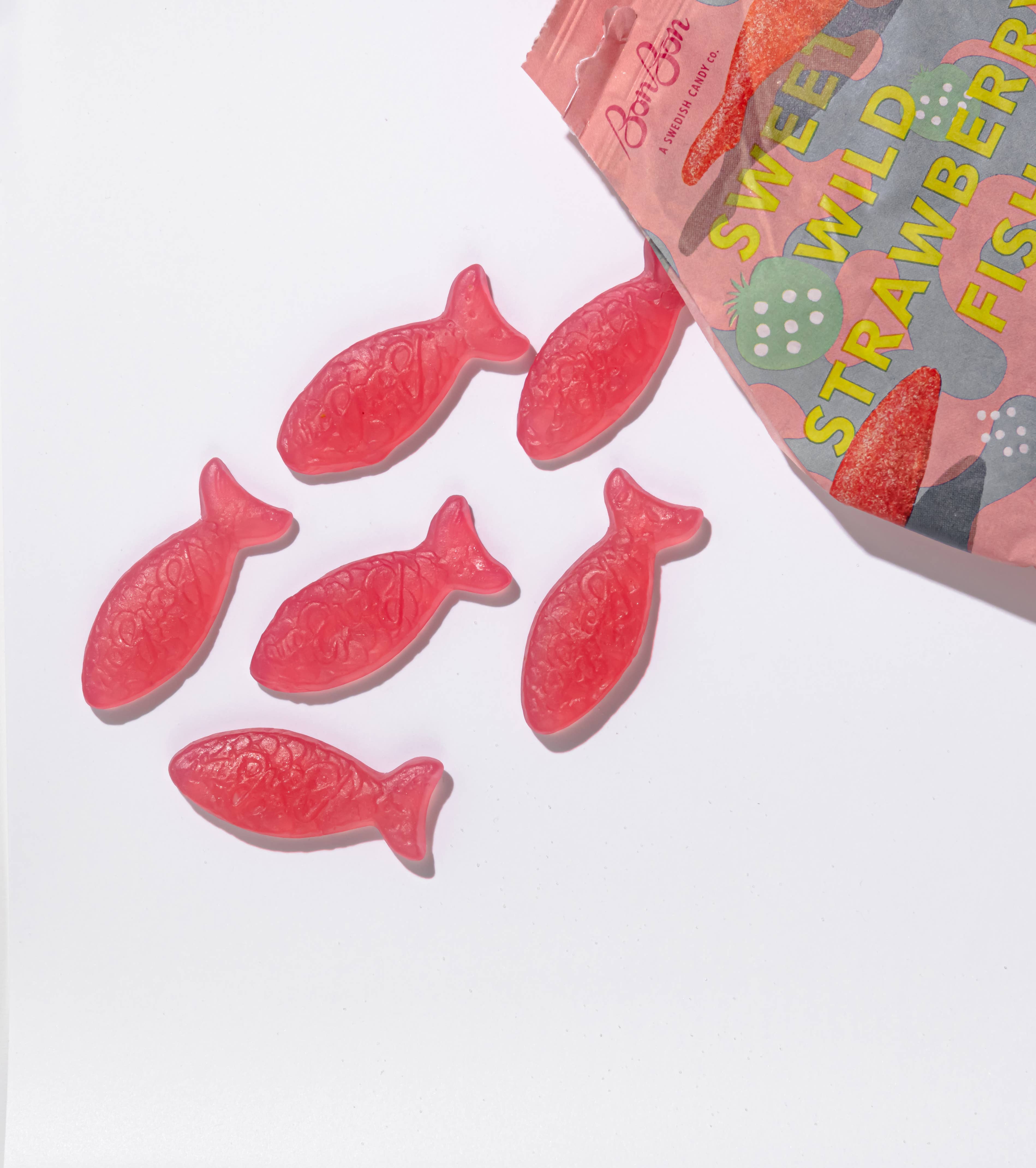 Six pink fish-shaped gummy candies are scattered near an open bag labeled Candy: BonBon - Sweet Wild Strawberry Fish (150g), a delightful Swedish candy that's perfect for those seeking vegan and gluten-free treats.