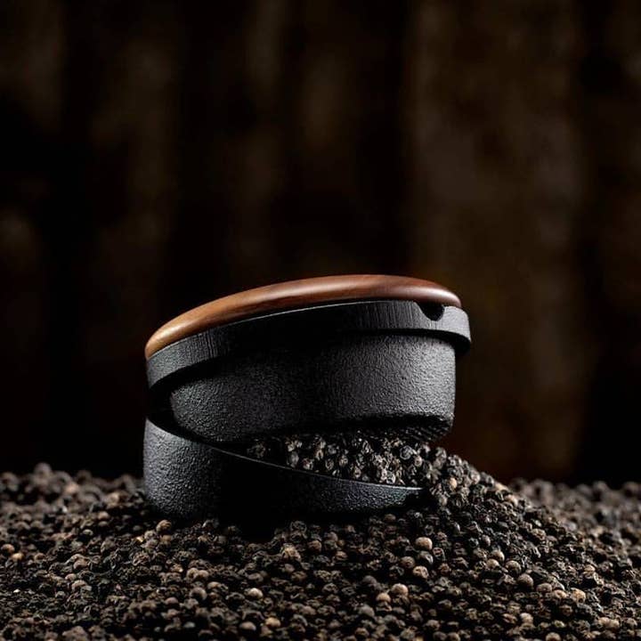 The Skeppshult Swing Spice Mill with Walnut Lid, crafted from cast iron and topped with rich walnut, is displayed on a bed of whole black peppercorns against a dark backdrop.