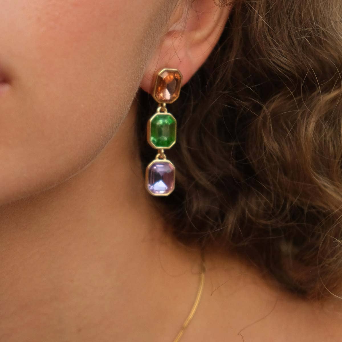 A close-up of an ear with Multi-Colored Crystal Dangle Earrings showcases three emerald cut glass stones in brown, green, and purple. Curly hair is partially visible, accentuated by elegant gold backings.