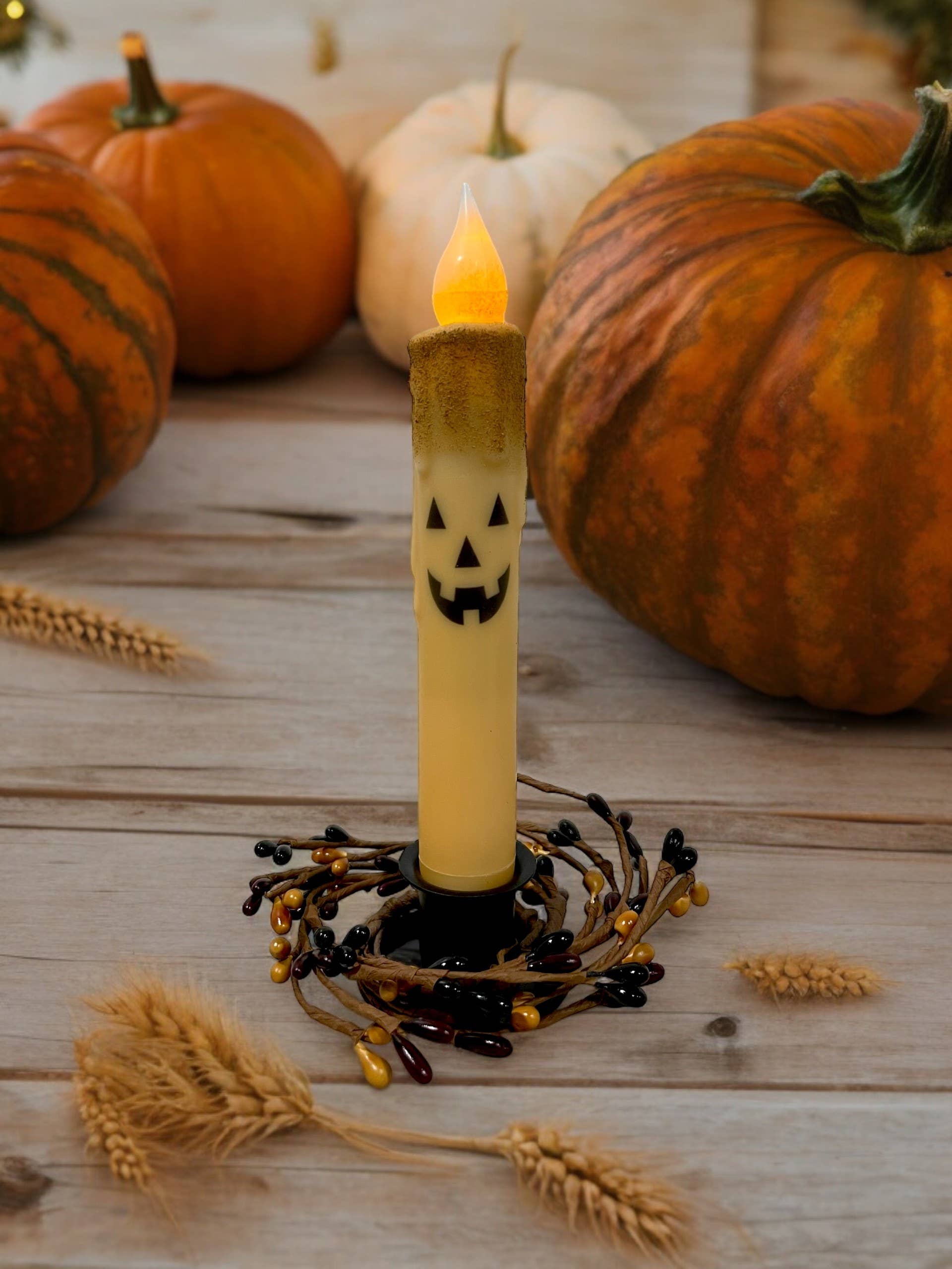 An enchanting glow emanates from the Jack-O-Lantern Halloween Battery Operated Timer Taper Candle in orange, surrounded by pumpkins and wheat on a wooden surface.